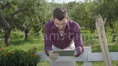 年轻的农<strong>民用</strong>平板电脑站在篱笆后面的前景，夏天的花园在背景。 那个人